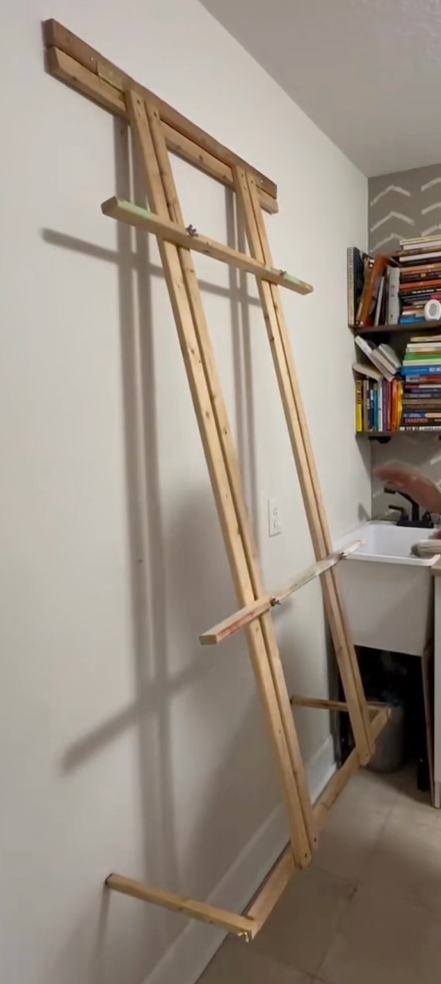A wooden leaning rack or easel made from natural wood planks, leaning against a white wall in an indoor room with shelves and a sink visible in the background.
